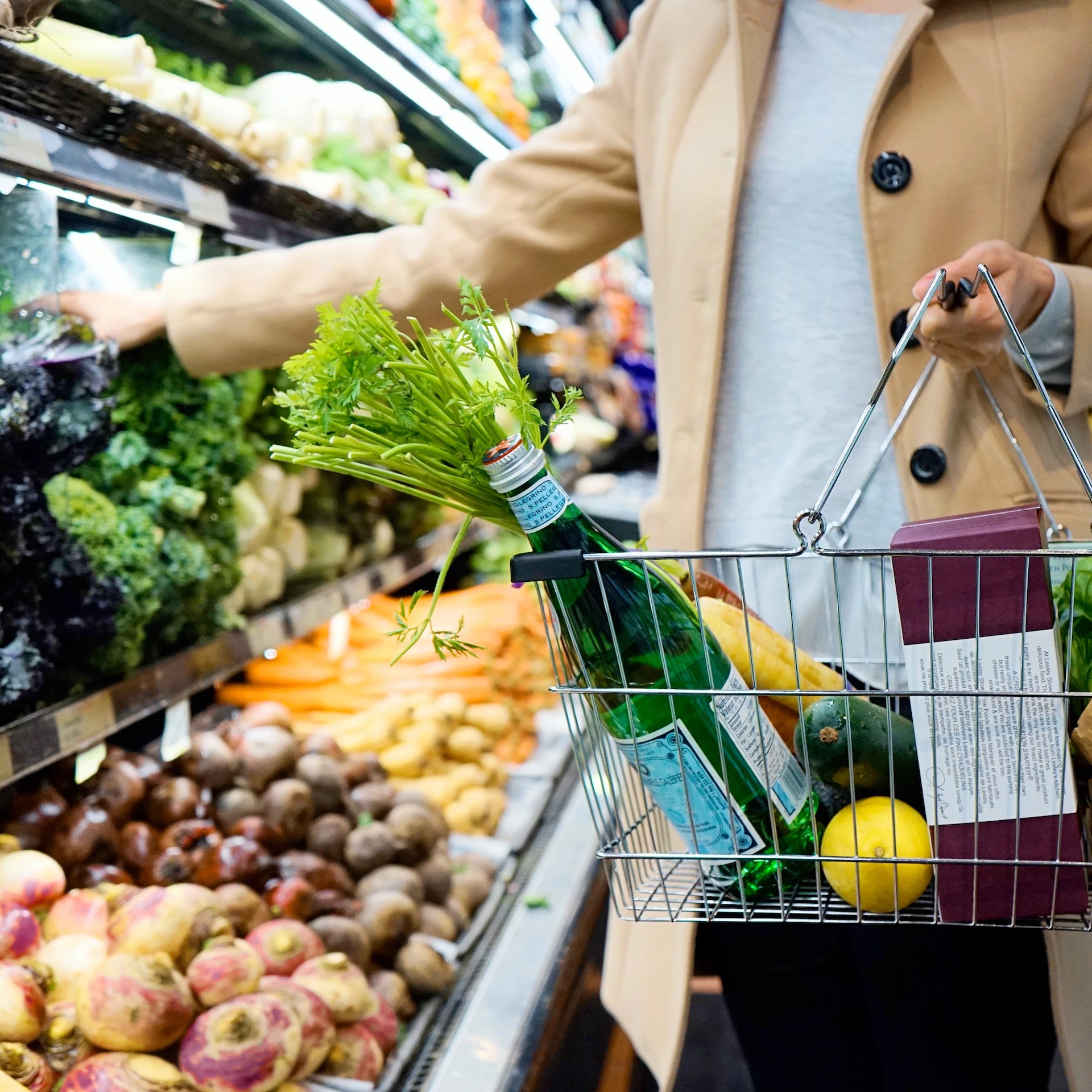 woman wearing camel coloured coat holding a shopping basket and reaching for vegetables