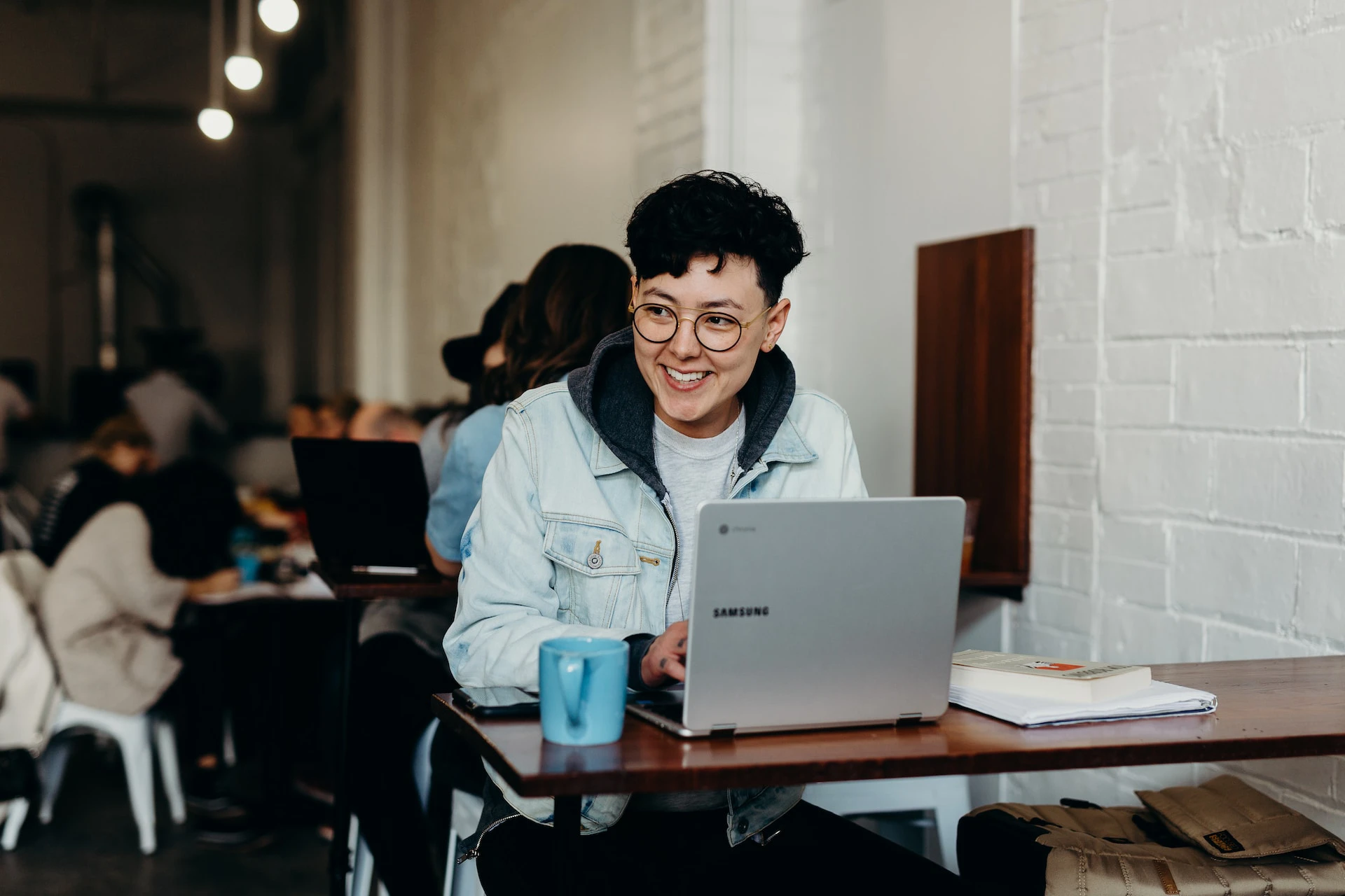 person with short dark hair and glasses sitting in a cafe working at a laptop and smiling